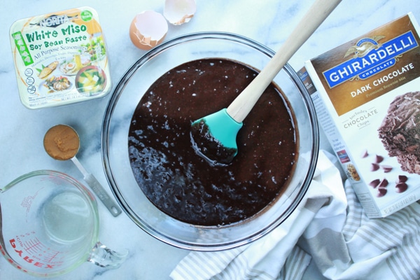 Brownie mix in a glass bowl on a white surface with a box of white miso paste on the side