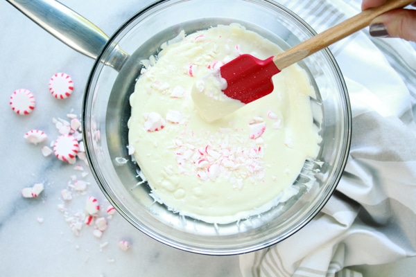 A glass mixing bowl filled with melted white chocolate with chunks of peppermint candies on top