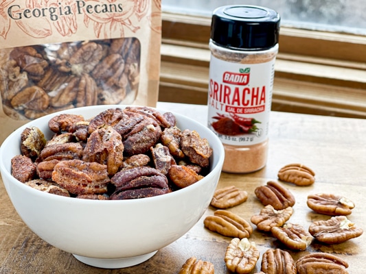 A white bowl filled with golden toasted spiced pecans on a wooden boards with a bag of pecans and sriracha seasoning behind it and loose pecans on the board.