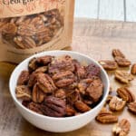 Spiced whole pecans in a white bowl with a bag of pecans and loose pecans on the side on a wooden board.