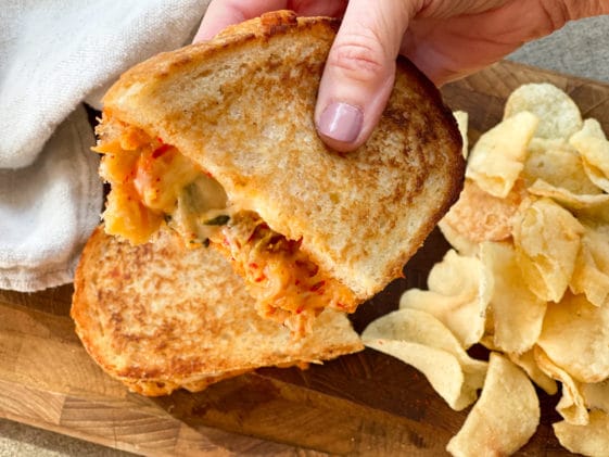 A woman's hand holing up half of a kimchi grilled cheese sandwich with potato chips on the side.