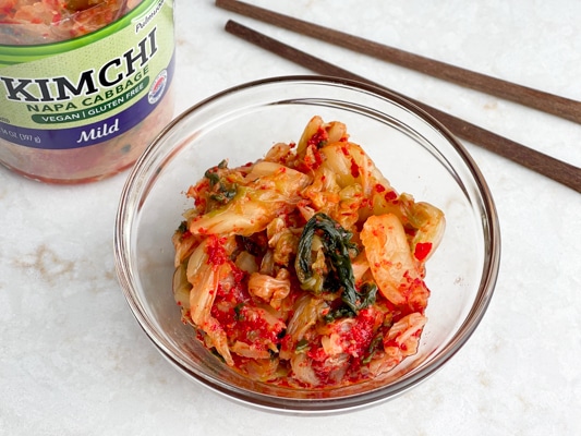 A clear bowl filled with kimchi on a white surface with a jar of kimchi and chopsticks behind it.