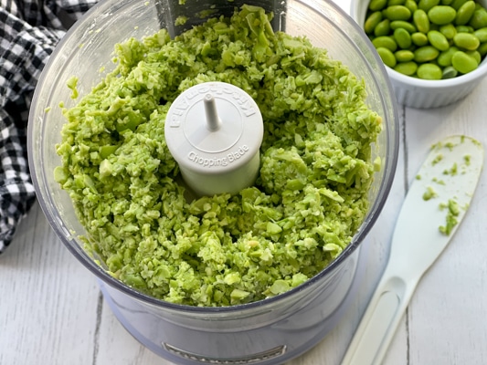 Edamame pulsed inside a food processor with a side of edamame pods and a white knife on the side.
