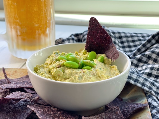 Goat cheese edamame dip in a small white bowl with edamame pods and blue corn tortilla chips on the side and a cold beer in a frosty mug behind.