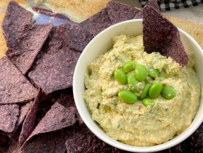 Goat cheese edamame dip in a small white bowl with edamame pods and blue corn tortilla chips on the side.