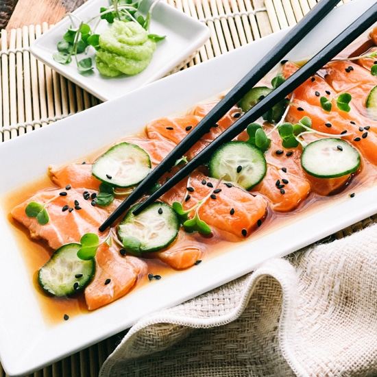 Slices of fresh sashimi salmon on a white platter with sliced cucumbers on top and sprinkles of black sesame seeds, with black chopsticks and a small white bowl of green wasabi on the side..