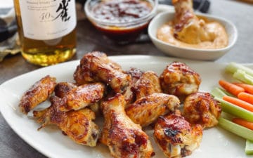 A bottle of Japanese whiskey placed behind a white plate of saucy chicken wings, celery and carrot sticks, with a bowl of aioli dip and extra sauce behind.