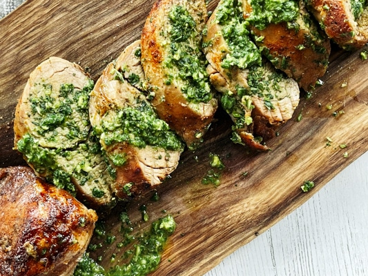 Thick slices of pork tenderloin on a rustic wooden board topped with a vibrant green cilantro, mint, basil sauce.