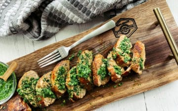 Thick slices of pork tenderloin on a rustic wooden board topped with a vibrant green cilantro, mint, basil sauce and a small bowl of sauce and a fork on the side.