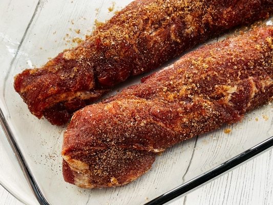 Two raw pork tenderloins rubbed with spices in a glass container on top of a white board.