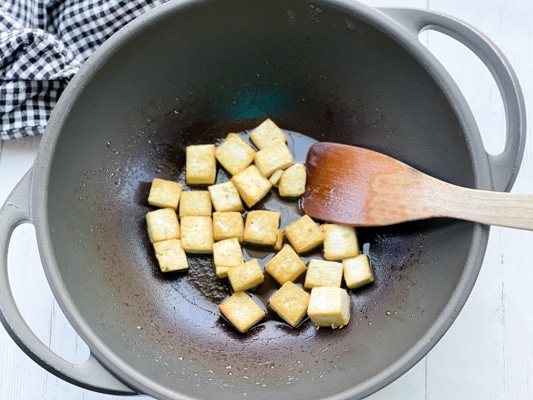 Tofu cubes being fried in a large wok with a wooden spatula.