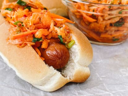 Close up of a hot dog in a bun topped with kimchi slaw with extra slaw on the side in a clear glass bowl.