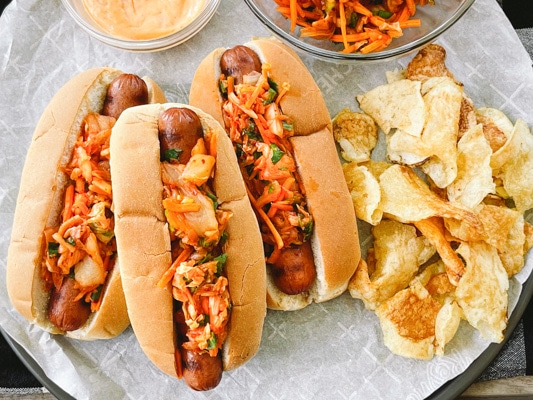 Three hot dogs in buns topped with kimchi slaw with potato chips, extra slaw and aioli on the side.