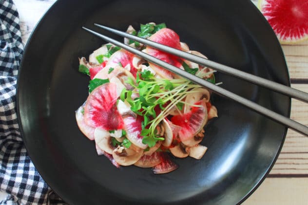 A vibrant mushroom and radish salad topped with sprouts inside a black bowl, with silver chopsticks on the side.