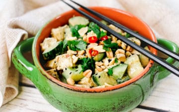 A green bowl filled with a vibrant spicy Asian tofu salad with cucumbers, cilantro and peanuts, and black chopsticks on top.