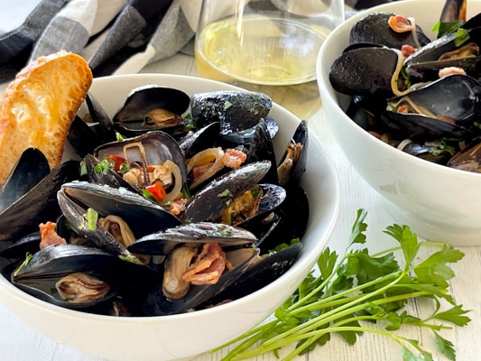 Two white bowls filled with steamed mussels and crusty bread on a white board, with a glass of white wine and green herbs on the side.