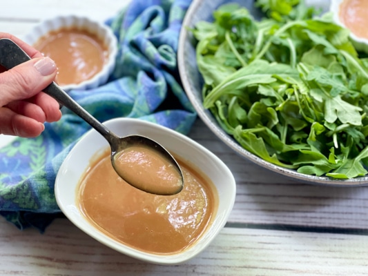 A woman's hand holding a spoon over a white bowl of homemade miso dressing with a bowl of greens and a blue napkin in the background on a white board.