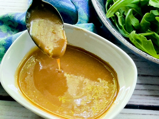 A spoon inserted into a white bowl of homemade miso dressing with a bowl of greens in the background on a white board.