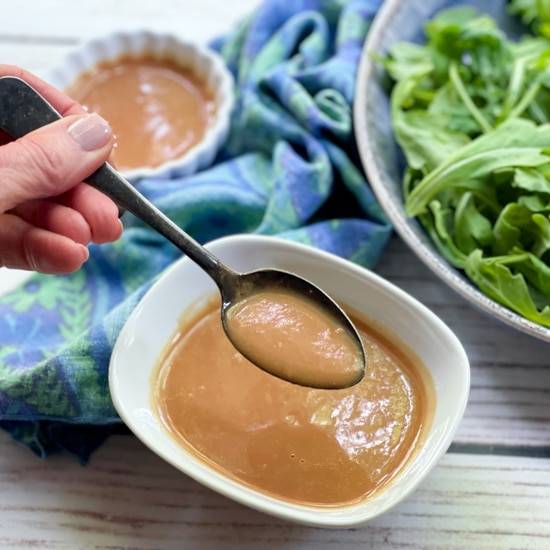 A woman's hand holding a spoon over a white bowl of homemade miso dressing with a bowl of greens and a blue napkin in the background on a white board.