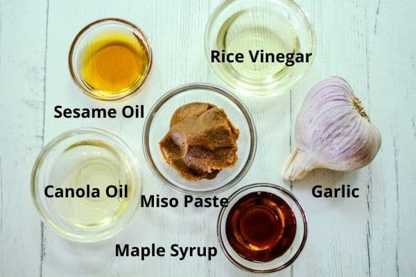 Ingredients for making miso dressing on a white board including rice vinegar, maple syrup, garlic, miso paste, sesame oil, and canola oil.