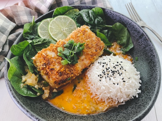 A beautiful piece of golden crusted halibut fillet in a black bowl with a vibrant red curry sauce at the bottom of the bowl, and white steamed rice and spinach on the side.