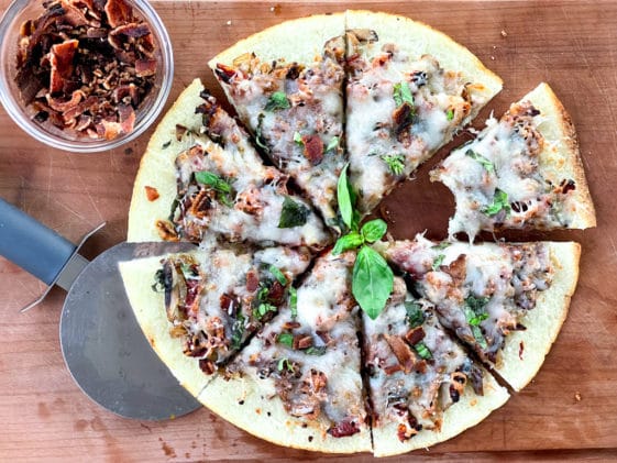 A sliced cauliflower pizza pie topped with bacon, shiitake mushrooms, basil, and melty cheese, with bacon bits on the side and a pizza cutter, on top of a wooden board.