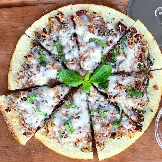 A sliced cauliflower pizza pie topped with crispy bacon, shiitake mushrooms, basil, and melty cheese, with a pizza cutter on top of a wooden board.