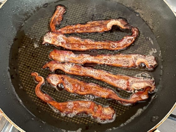 Several slices of bacon frying up in a black frying pan.