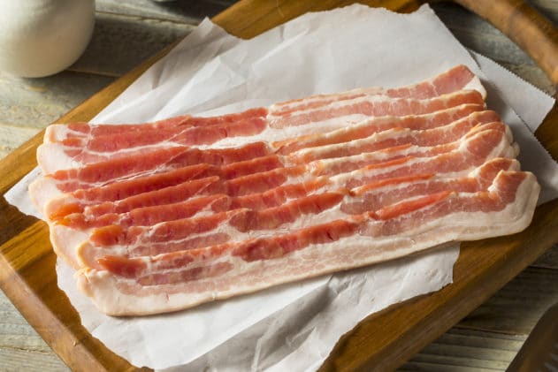 Several slices of raw bacon on top of parchment paper on a wooden board.