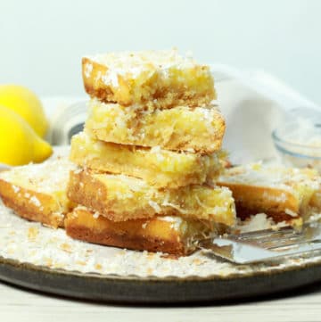 A stack of gluten-free lemongrass coconut squares on a white plate with fresh lemons in the background.