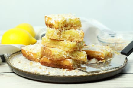A stack of gluten-free lemongrass coconut squares on a white plate with fresh lemons in the background.