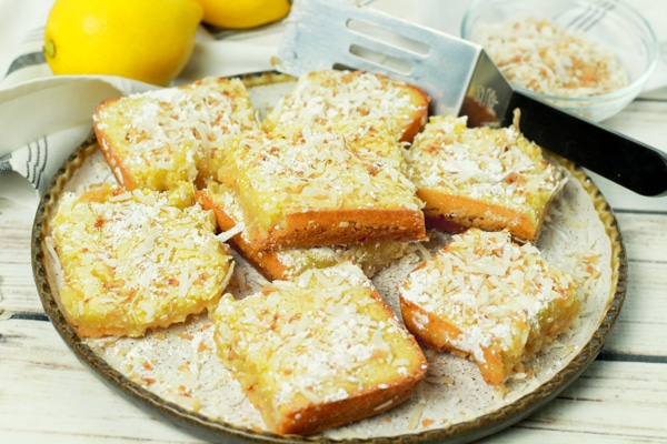 Gluten-free lemongrass coconut bars topped with toasted coconut flakes and powdered sugar on a white plate with a spatula and fresh lemons on the side.