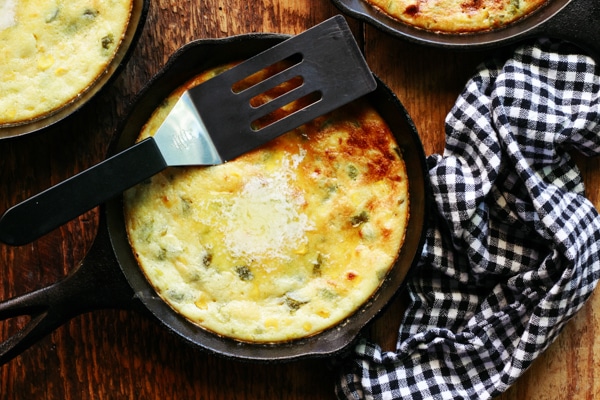 A baked jalapeño corn bread in a mini skillet with a spatula on top and a black and white checkered napkin on the side, and two other mini skillets behind, on top of a wooden board.