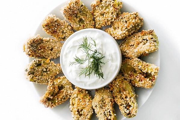 Crispy air-fried artichoke hearts placed in a circle on a white plate with a small bowl of yogurt dip in the middle. A yummy holiday appetizer.