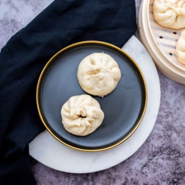 Two Chinese steamed pork buns on a round black plate with a bamboo steamer on the side filled with steamed buns, on top of a gray marbled surface
