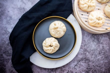 Two Chinese steamed pork buns on a round black plate with a bamboo steamer on the side filled with steamed buns, on top of a gray marbled surface