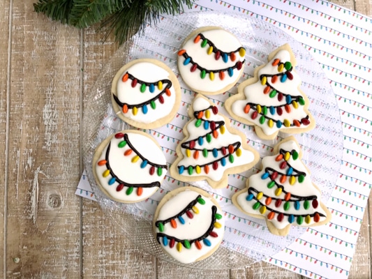 Decorated holiday sugar cookies decorated with white frosting and holiday lights on top of a wooden board.