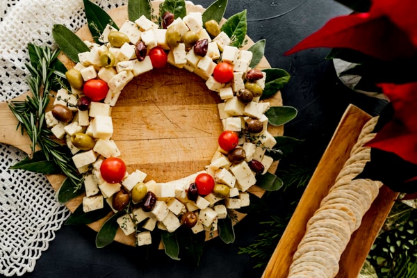 A round Christmas wreath cheese board with cubes of cheese with cherry tomatoes and olives on leaves with a basket of crackers on the side.