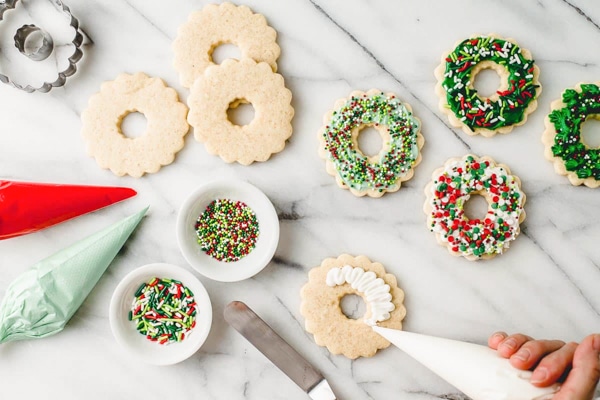 A hand piping the frosting on top of a cut-out wreath-shaped sugar cookie with two small bowls of colorful sprinkles and extra piping bags on the side, all on top of a marble surface.