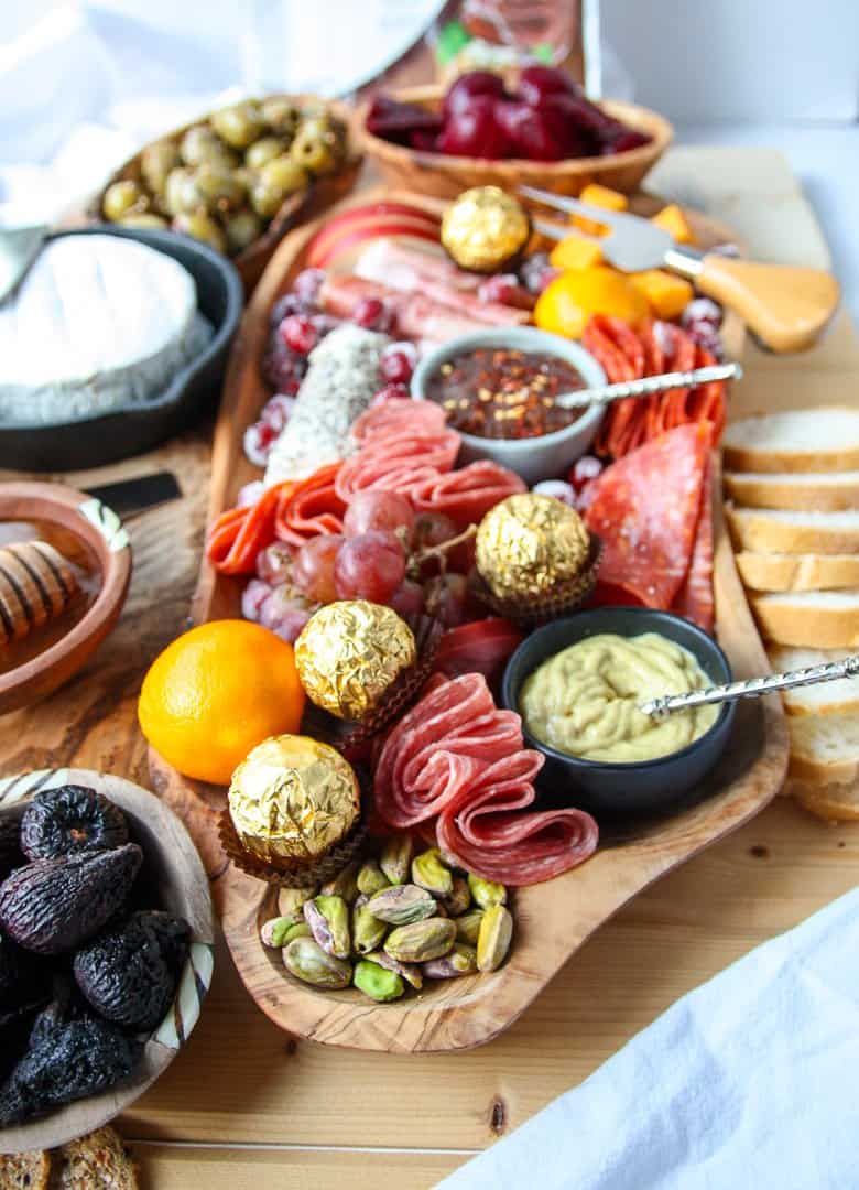 A large wooden board topped with meats and cheeses, olives and nuts in this magnificent holiday charcuterie board with bread slices, crackers, and dips on the side.