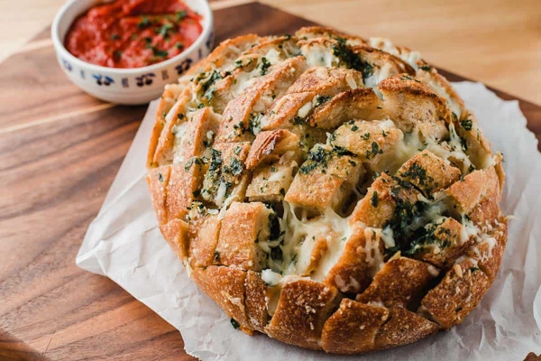 A beautiful loaf of pull-apart cheese bread on a wooden board with a red dipping sauce on the side.