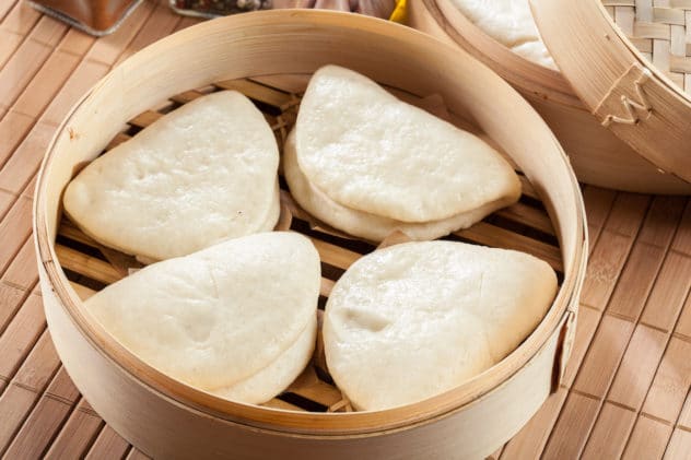 Fully white bao buns steamed in an open-lid bamboo steamer on top of a bamboo place mat.
