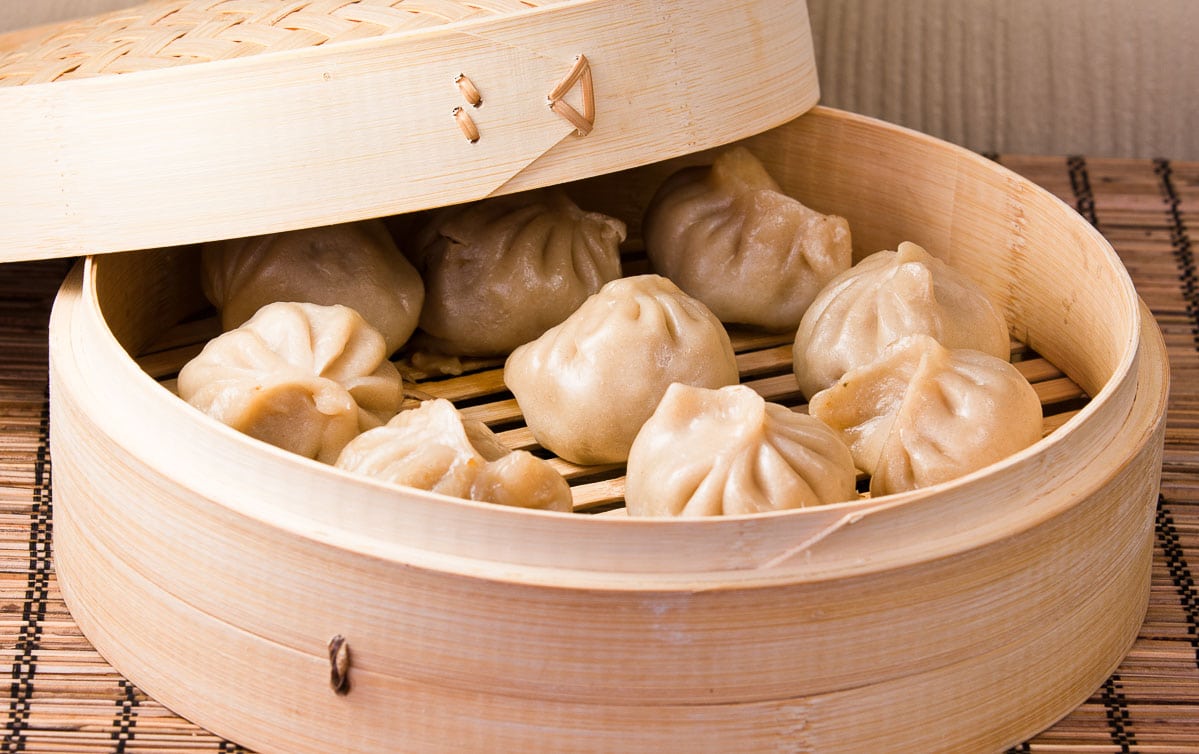 Steamed Chinese pork dumplings in a bamboo steamer with the lid on the side, on top of a bamboo place mat.