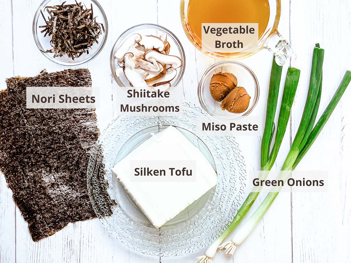 Labeled ingredients for making miso soup on top of a white wooden board including miso paste, shiitake mushrooms, silken tofu, nori sheets, vegetable broth, and green onions.