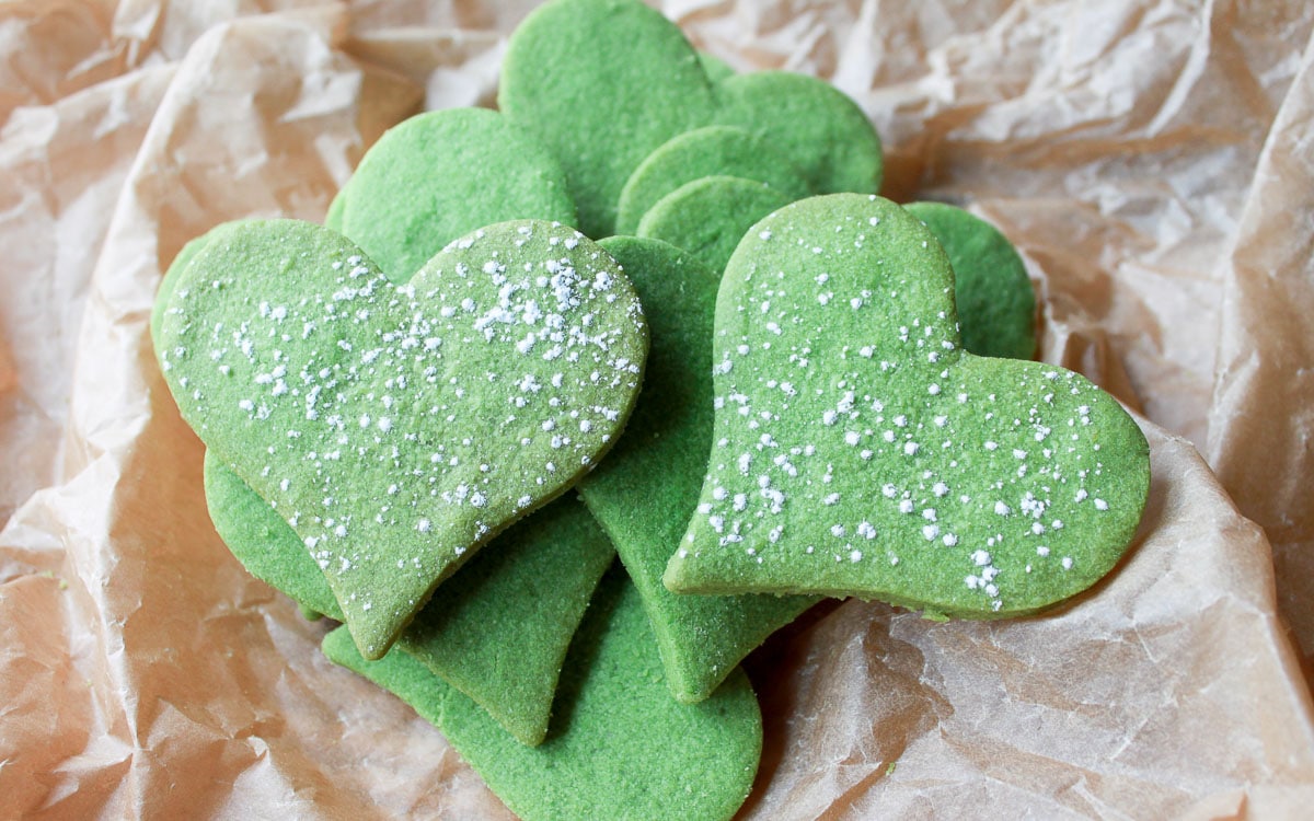 Heart-shaped matcha sugar cookies sprinkled with powdered sugar piled on top of brown parchment paper.