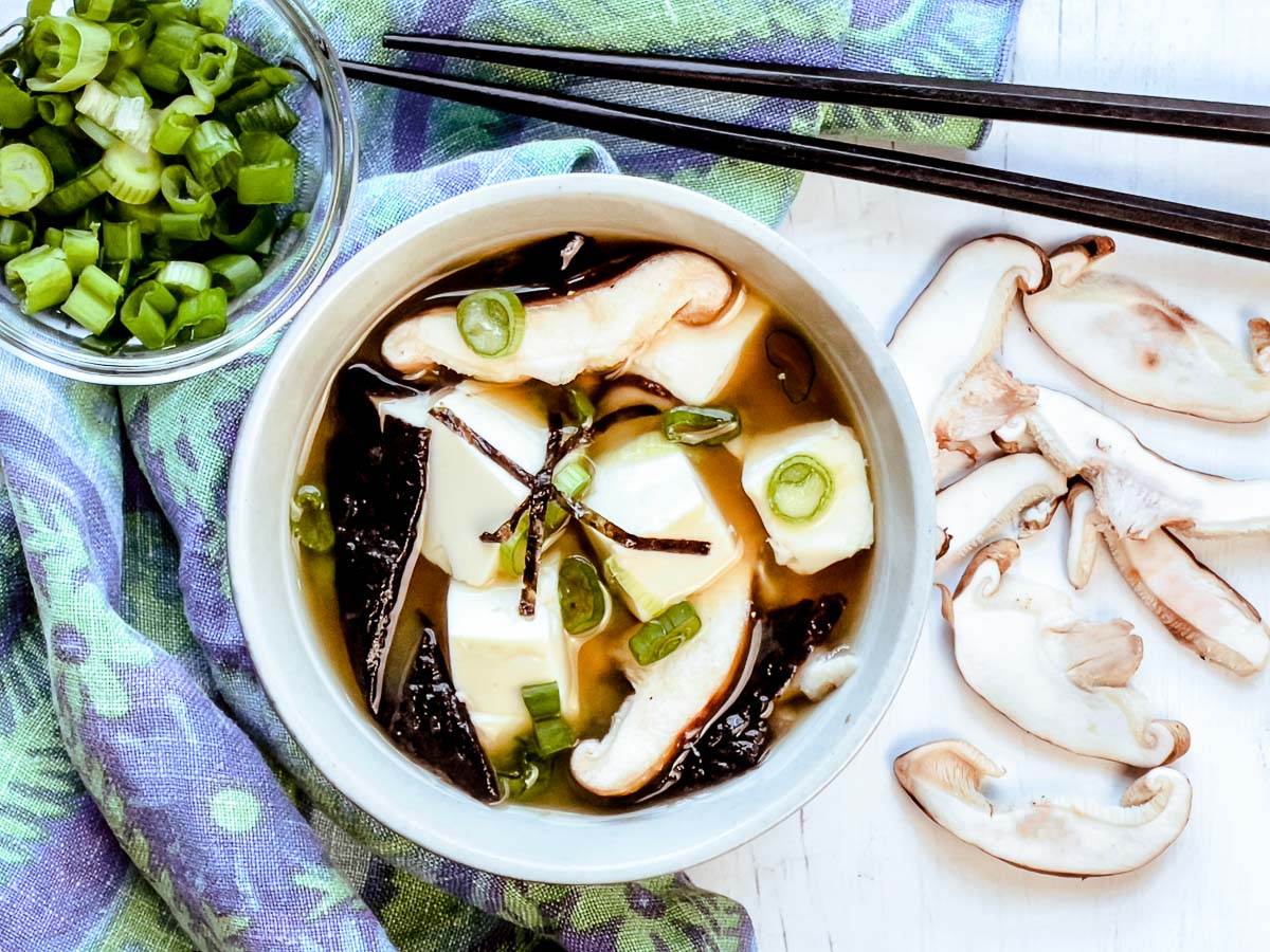 A white bowl filled with miso soup with tofu and shiitake mushrooms on top of a printed blue napkin with shiitake mushrooms and chopped green onions, and black chopsticks on the side.