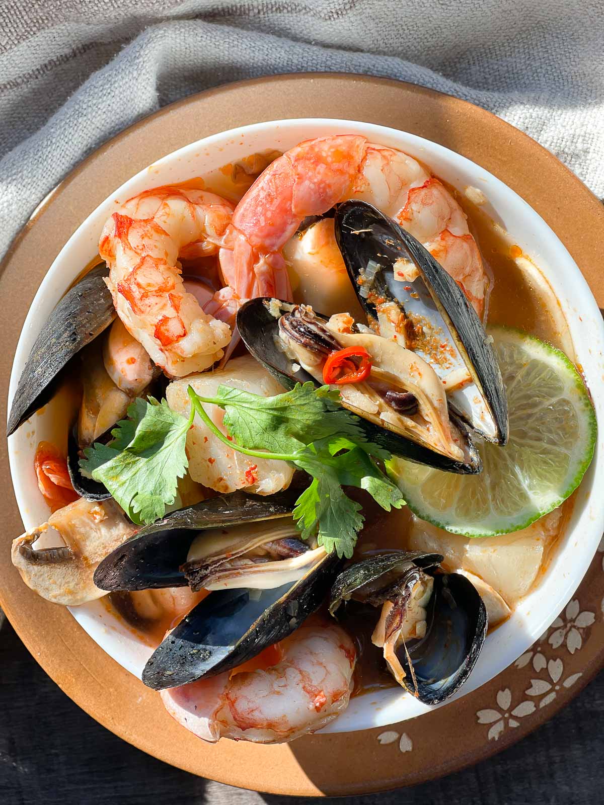 A white bowl of Thai Coconut Curry Seafood Soup with shrimp, mussels and lime on top of a rust-colored plate with fresh herbs on top placed on top of a linen napkin.