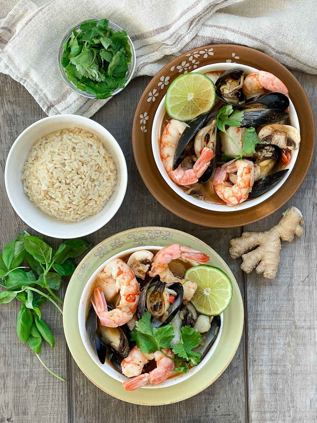 Two white bowls of Thai Coconut Curry Seafood Soup with shrimp, mussels and lime on top of a yellow and rust-colored plate plate with fresh herbs and a small bowl of rice on the side placed on top of a wooden board.