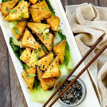 Golden-brown, crispy tofu steaks piled on top of cooked baby bok choy and sprinkled with seasonings plated on top of a white serving platter with small bowl of sauce and furikake seasoning on the side, and a pair of chopsticks on top and a napkin on the side.