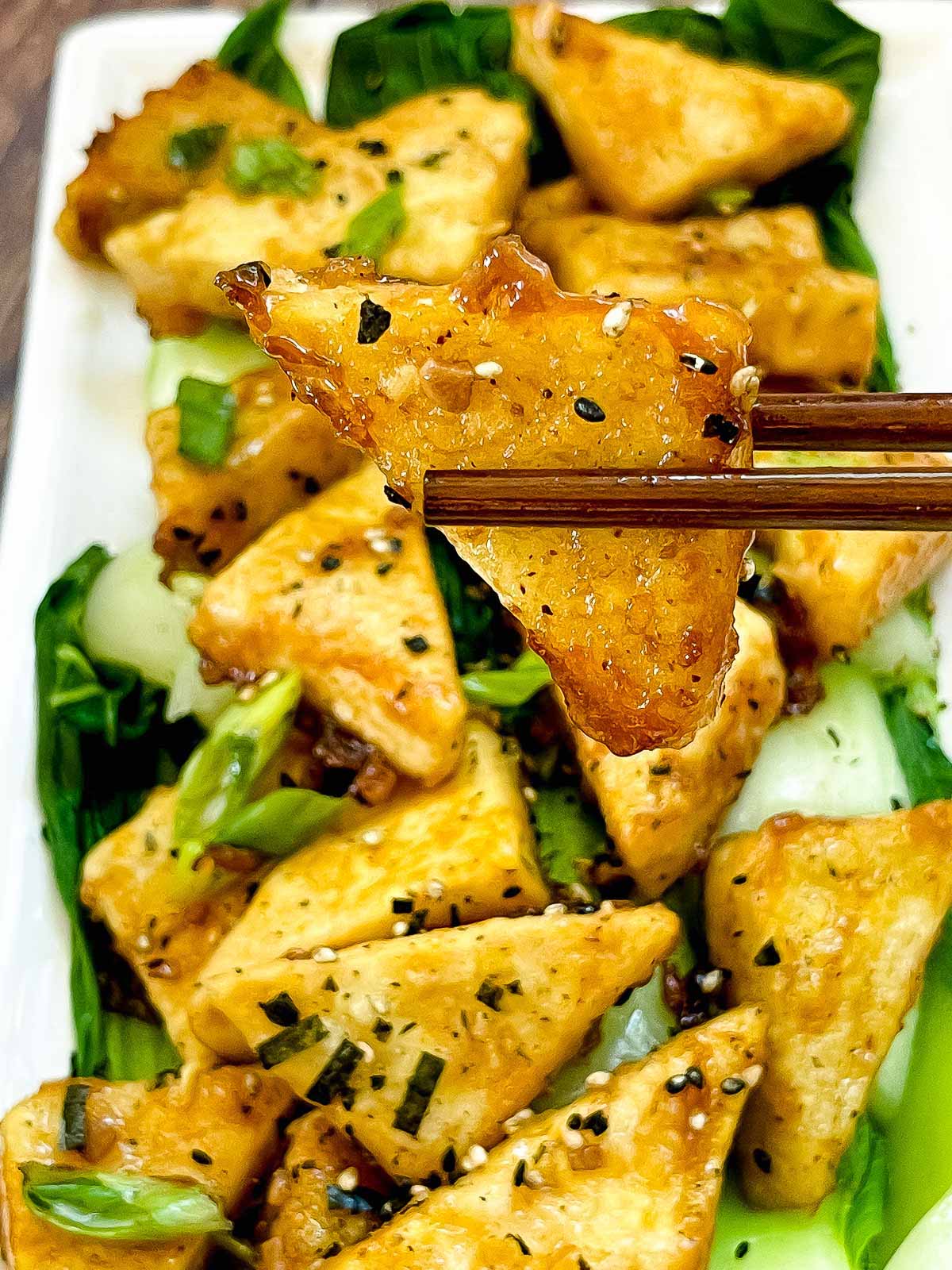 Golden-brown, crispy tofu steaks piled on top of cooked baby bok choy with a pair up chopsticks holding up one piece of tofu.